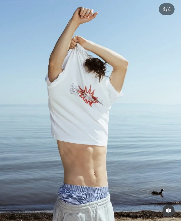 Ragazzo di spalle sulla spiaggia che si sfila la t-shirt RITMO, mare sullo sfondo